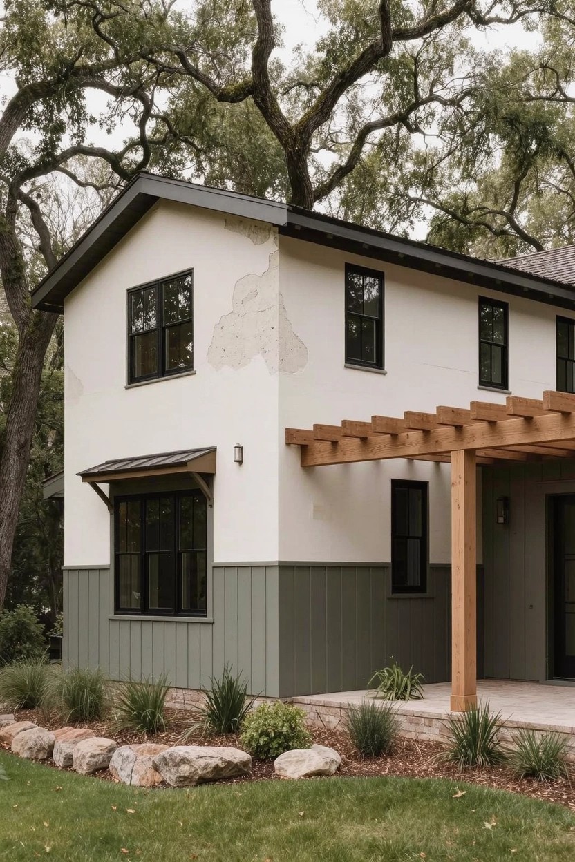 White upper siding and green lower siding on a gabled house with black windows, wooden pergola over side patio, oak trees, rocks, and grasses in yard.