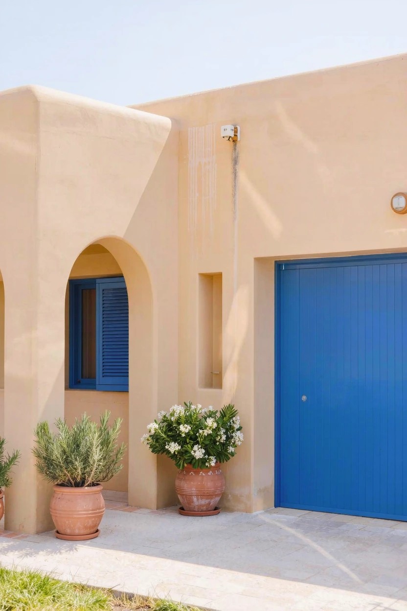Beige stucco house exterior featuring an arched window with blue shutters, a blue garage door, potted plants with white flowers and lavender bushes, and a stone pathway.