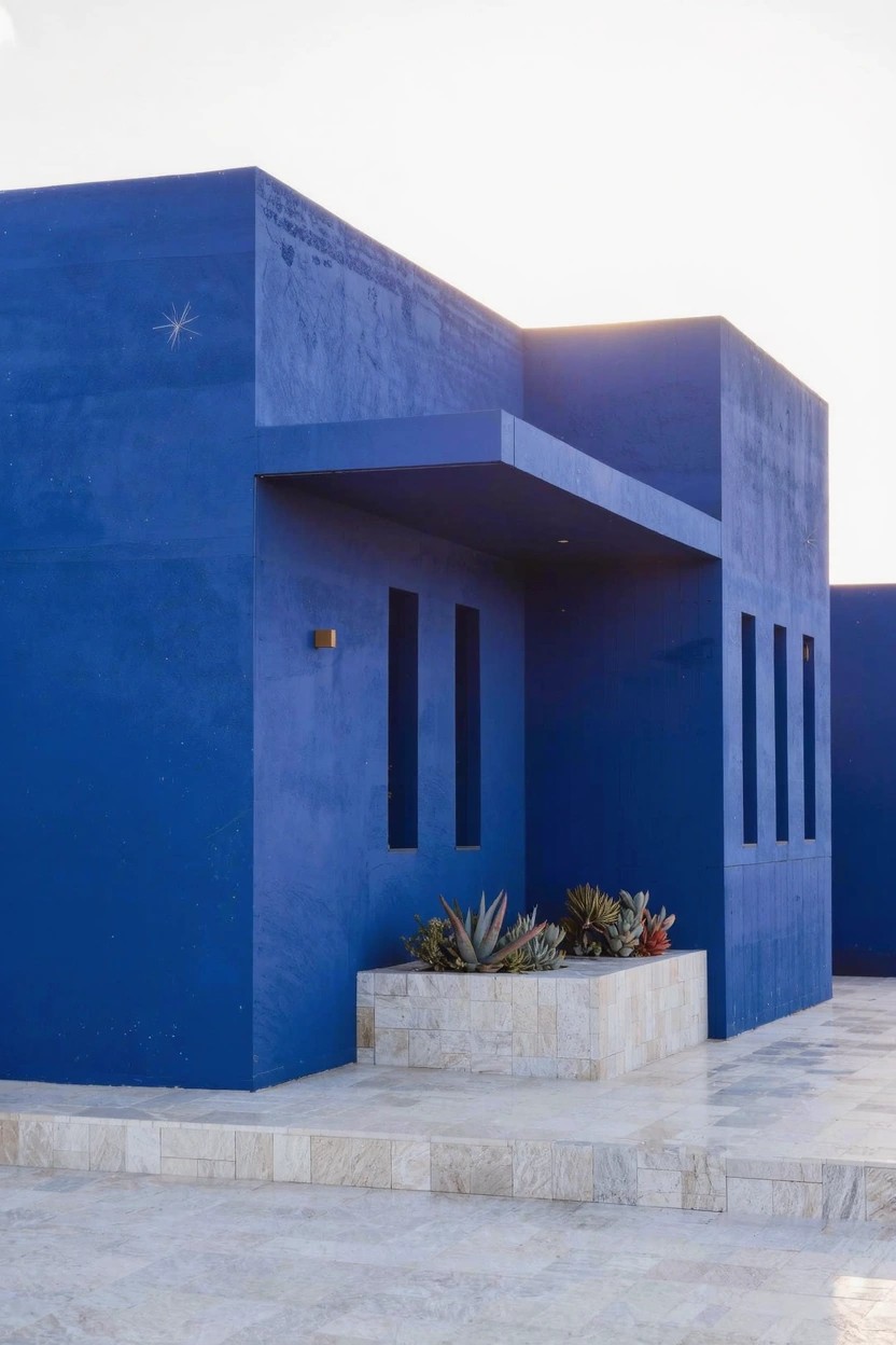 Boxy modern house exterior painted vibrant blue with narrow vertical windows, a projecting canopy over the entry, stone planter holding succulents, and light stone pavers in front.