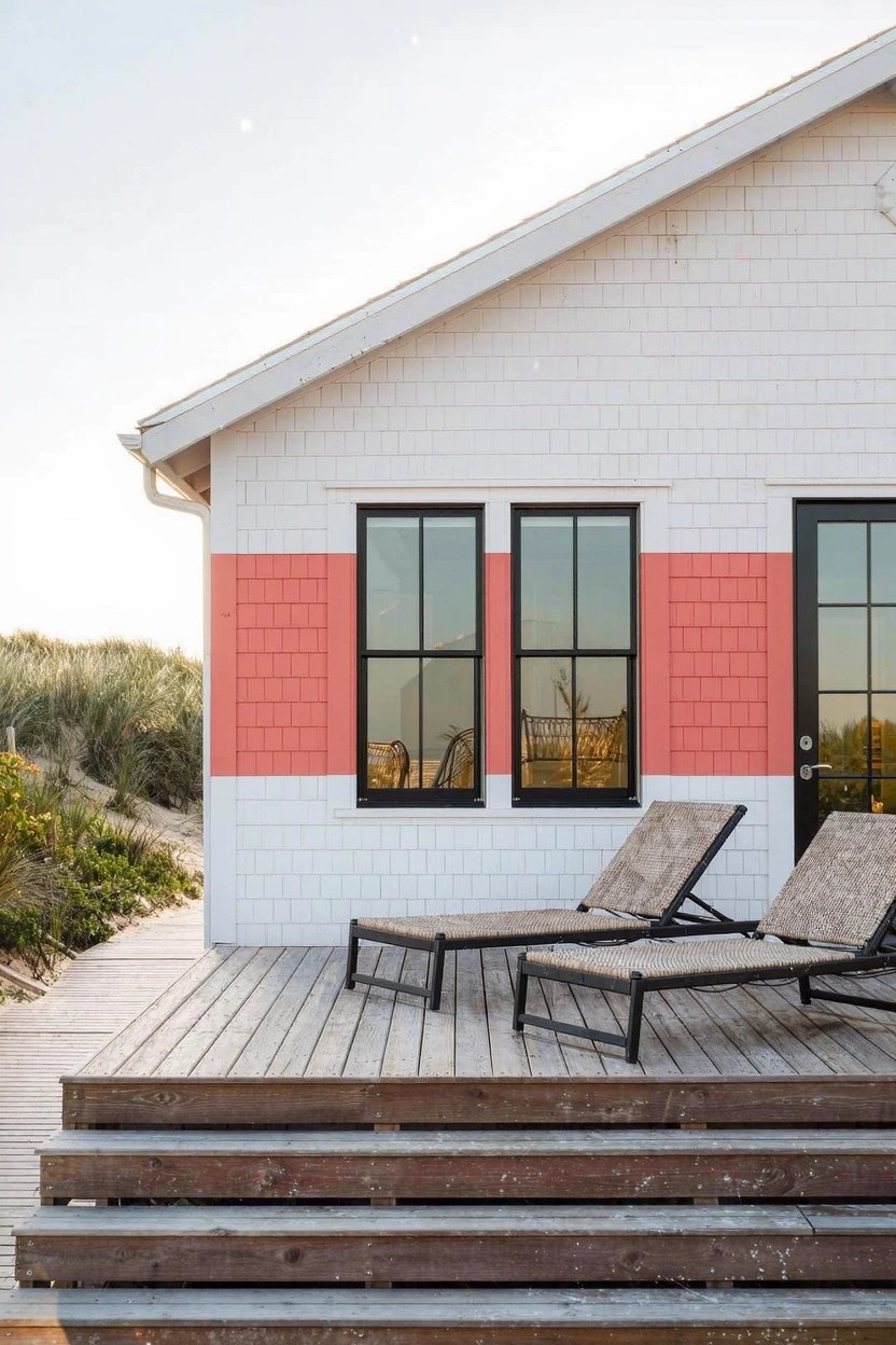 White and coral pink shingled house exterior with black-framed windows and glass door on a wooden deck with two lounge chairs, dunes and beach grass nearby at sunset.
