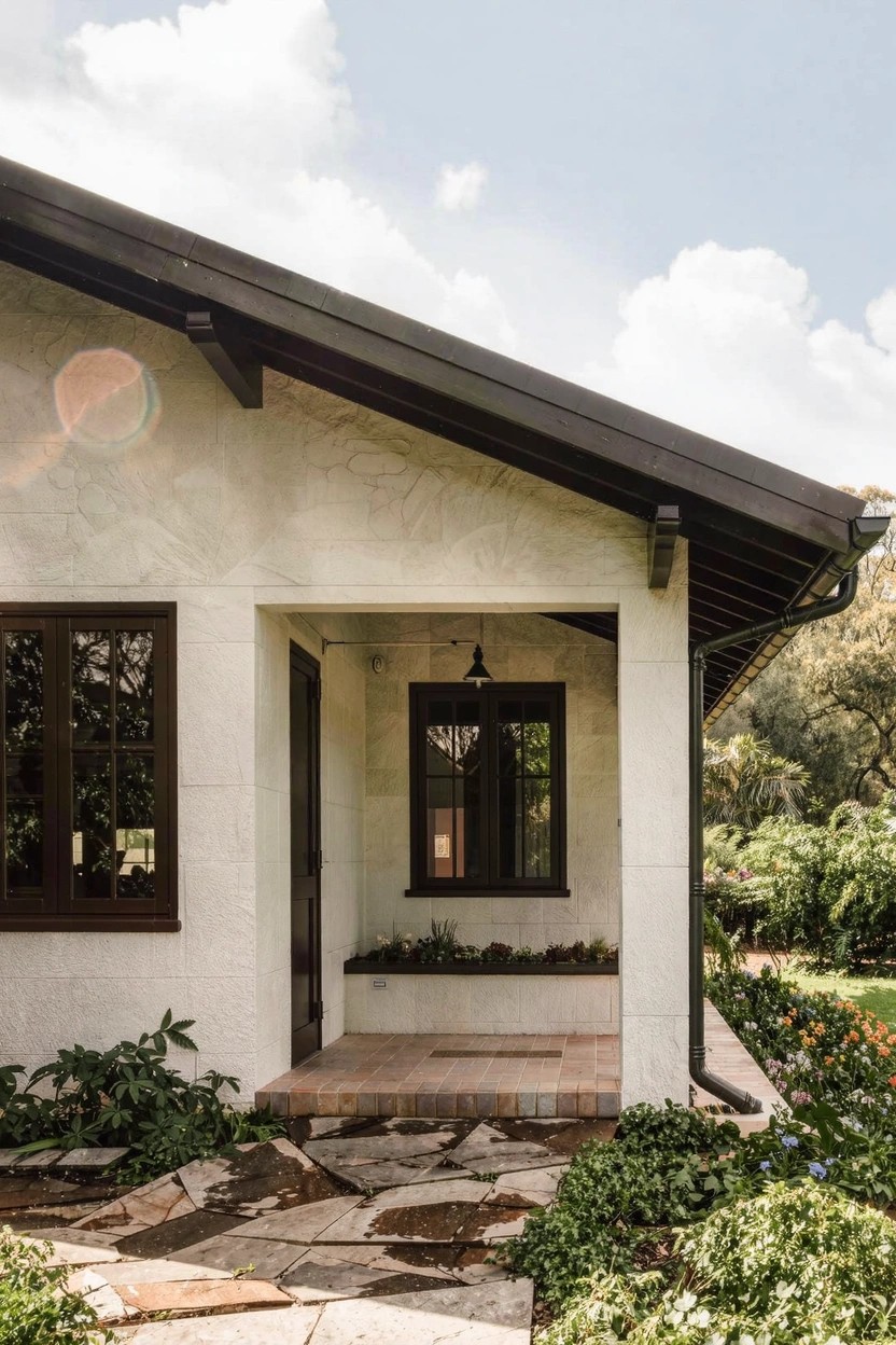 White stucco house corner with dark wood-framed windows and door, covered porch with hanging lantern, irregular stone steps leading to entry, planter box, and greenery around base under black roof eaves.