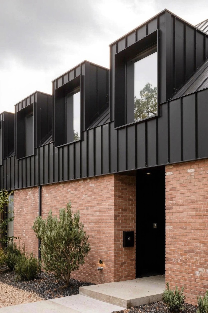 Modern house exterior featuring protruding black metal frames around upper windows over a red brick base wall, with a black front door, concrete steps, gravel ground cover, and small shrubs.