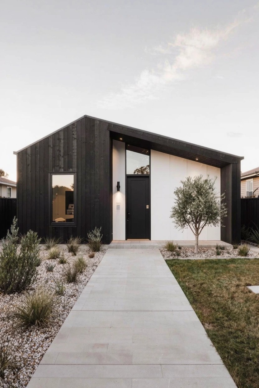 Modern single-story house with black vertical timber siding on most of the facade, white stucco on one side, angular overhanging roof, black front door, concrete paver pathway through gravel and native grasses, and a small olive tree nearby.