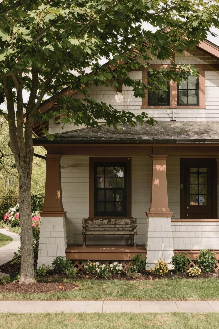 White Craftsman-style house with dark stained tapered porch columns, wooden bench swing, double windows, flower beds, and a large green tree beside a curved walkway.