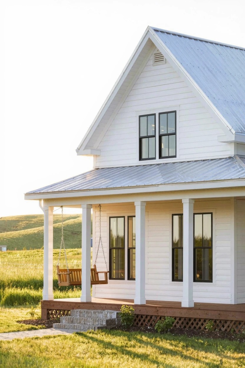 White board-and-batten sided house with gabled metal roof, black-framed windows, wraparound porch with columns and hanging swing, set in grassy field with distant hills.