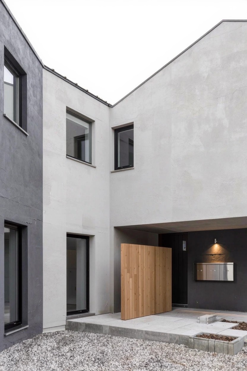 Corner view of a modern house exterior featuring dark gray plaster on one wall, light gray plaster on the adjacent wall, a vertical wooden entry panel and door, black-framed windows, and gravel paving.