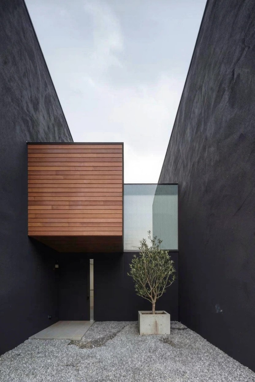 Tall matte black stucco walls form a narrow courtyard with gravel ground, a slim metal entry door, a cantilevered rectangular wooden box structure with horizontal slats and glass window above, and a small olive tree in a concrete planter.