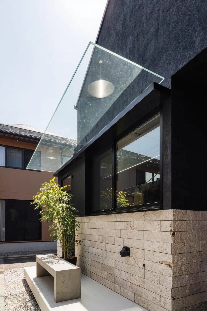 Modern house exterior with dark upper facade and sliding windows under a clear glass canopy with pendant light, light beige stone base, concrete bench, and potted bamboo plant beside pebbled ground.
