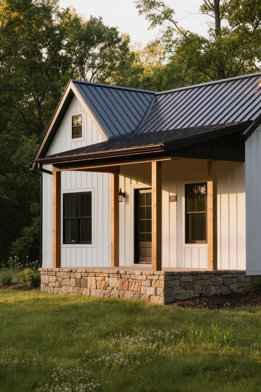 Small white board-and-batten sided house with dark standing seam metal gable roof, covered front porch on wooden posts, stone foundation, and surrounding grass and trees.