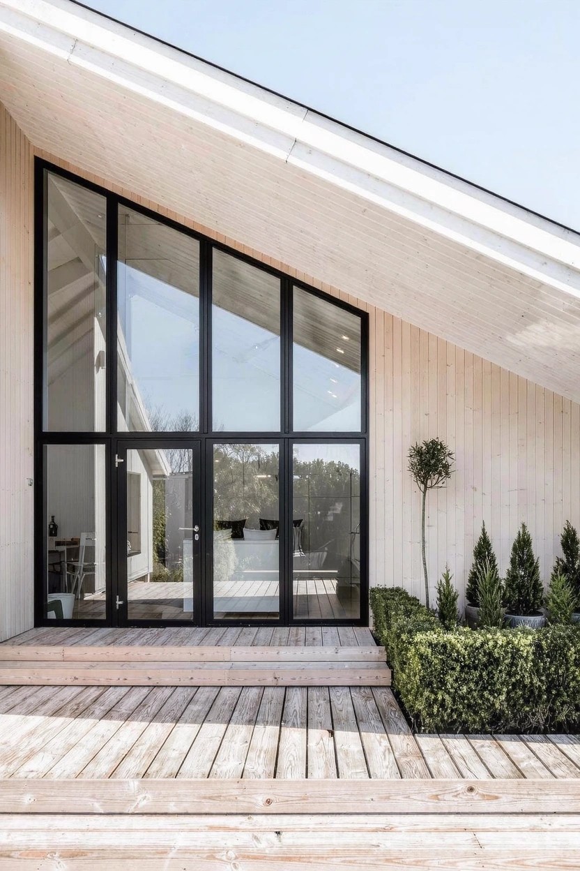 Modern small house with sloped light wood-clad roof over large black-framed glass entry doors and windows, wooden deck steps, and boxwood hedges in front.