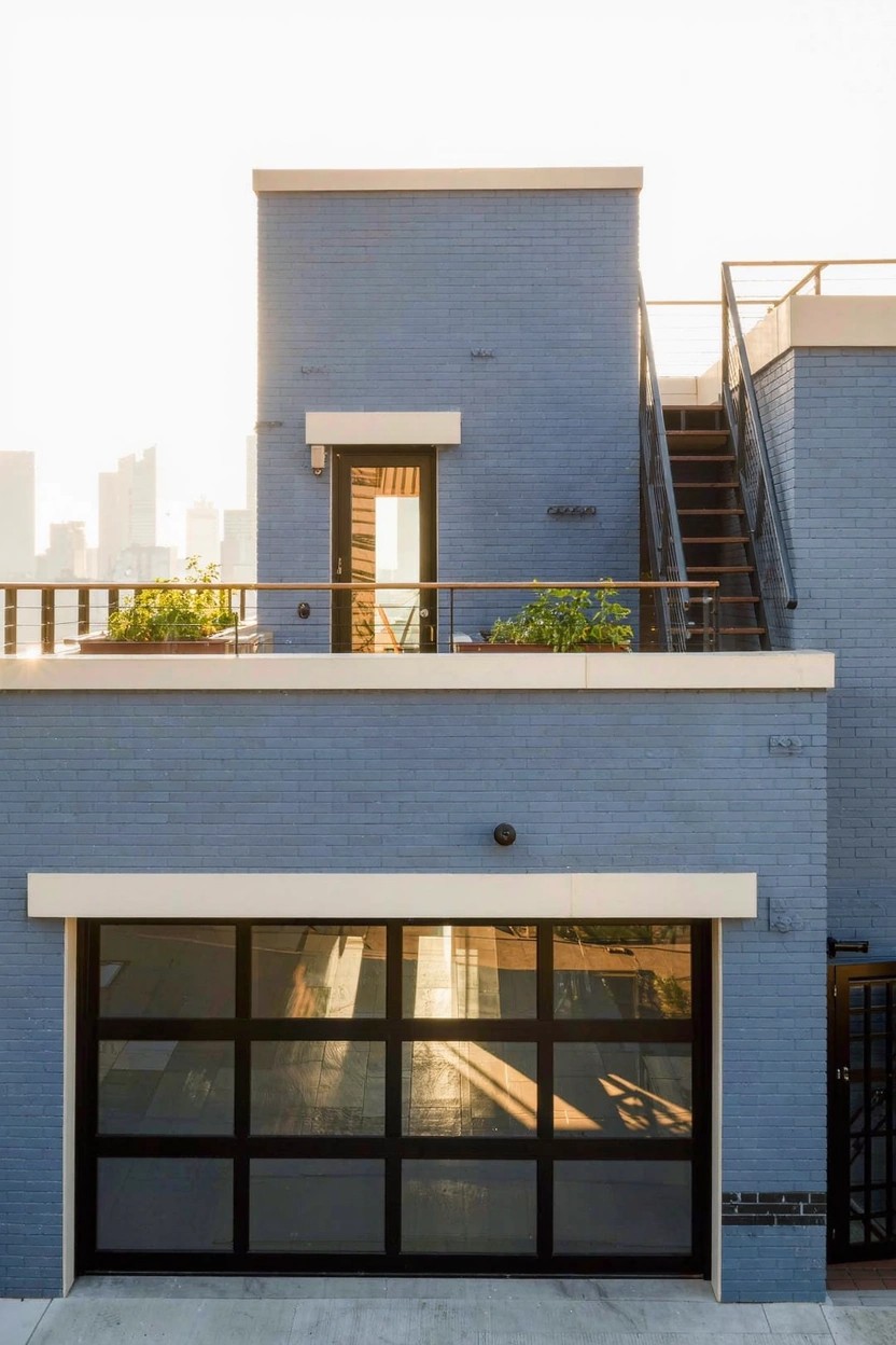 Modern two-story house exterior with soft blue brick walls, a large glass-paneled garage door, and a rooftop deck with metal stairs and potted plants against a city skyline at sunset.