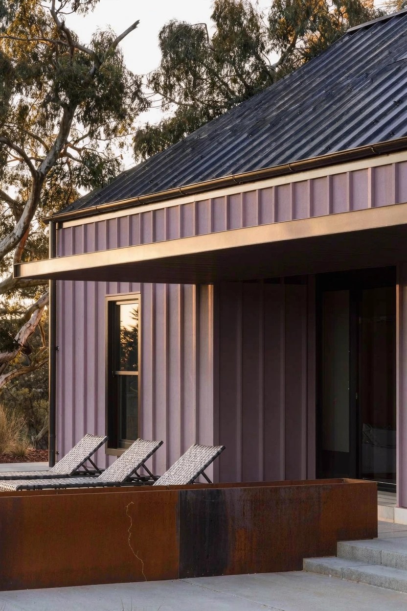 Modern house exterior with purple corrugated metal siding, black standing-seam roof, overhanging eaves, glass door and window, patio lounge chairs, corten steel raised planter bed, steps, and nearby eucalyptus trees in evening light.