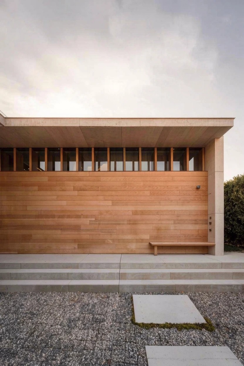 Modern house exterior featuring horizontal warm-toned wood cladding on the facade, concrete steps with a wood bench, gravel ground, and stone pavers beside greenery.