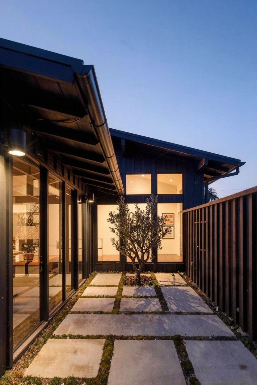 Black wood-clad modern house at dusk with wall lights on, large glass doors opening to a courtyard of square stone pavers around a small olive tree, wooden fence on one side.