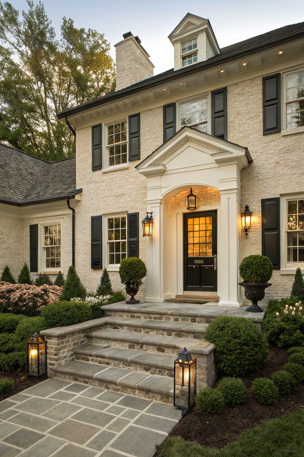 Two-story cream brick house exterior featuring a white pedimented portico around a black front door with glass panels, flanked by lanterns, black shutters on windows, stone steps, and boxwood shrubs in urns.