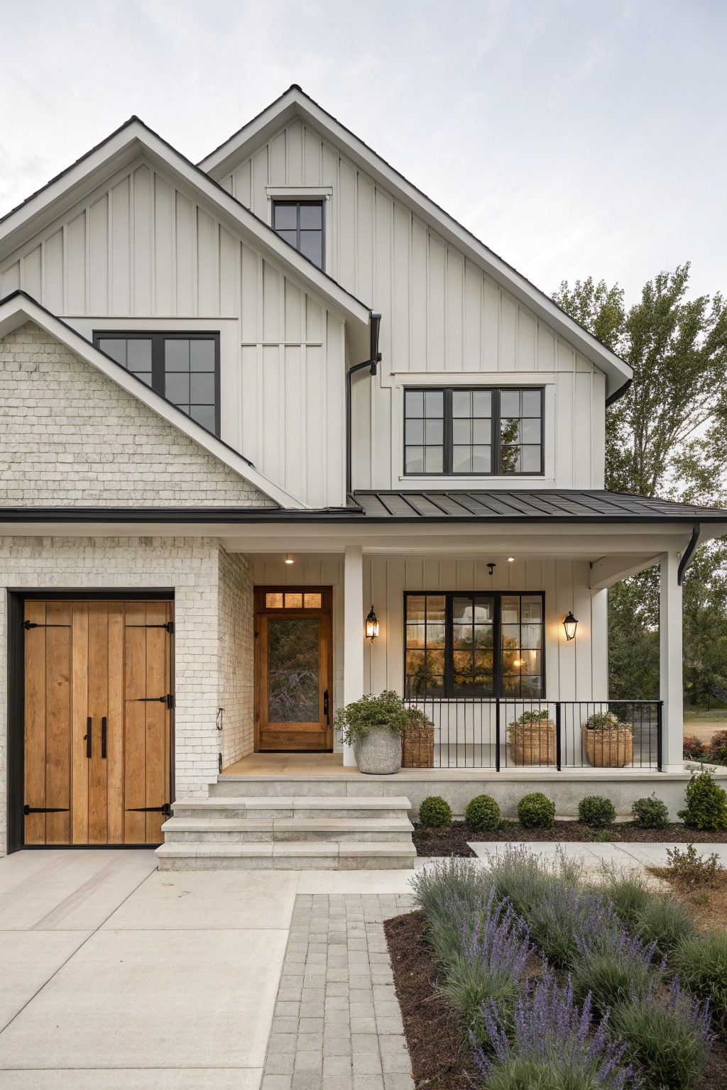 Two-story house exterior with cream brick on the base and garage, white board-and-batten siding above, black metal roof and window frames, wooden double garage doors, covered front porch with glass door, potted plants, and lavender landscaping along the driveway.