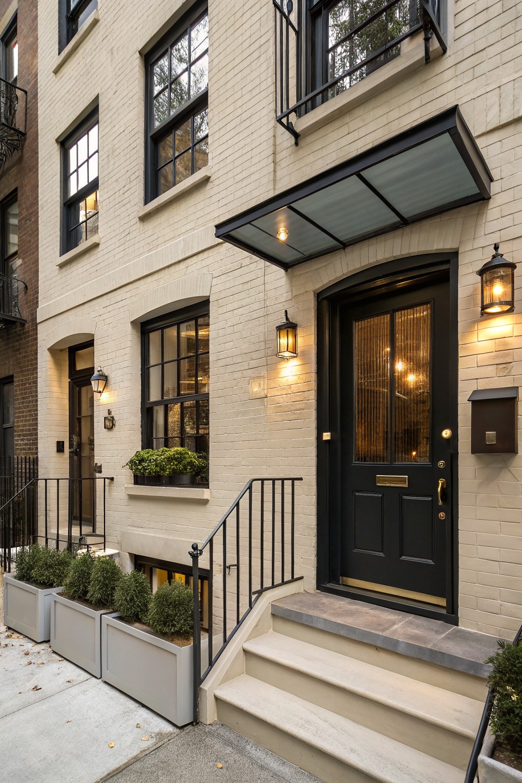 Cream painted brick townhouse exterior with black framed windows, arched black front door with glass panels, entry steps with black iron railing, potted boxwood shrubs, and lantern lights.