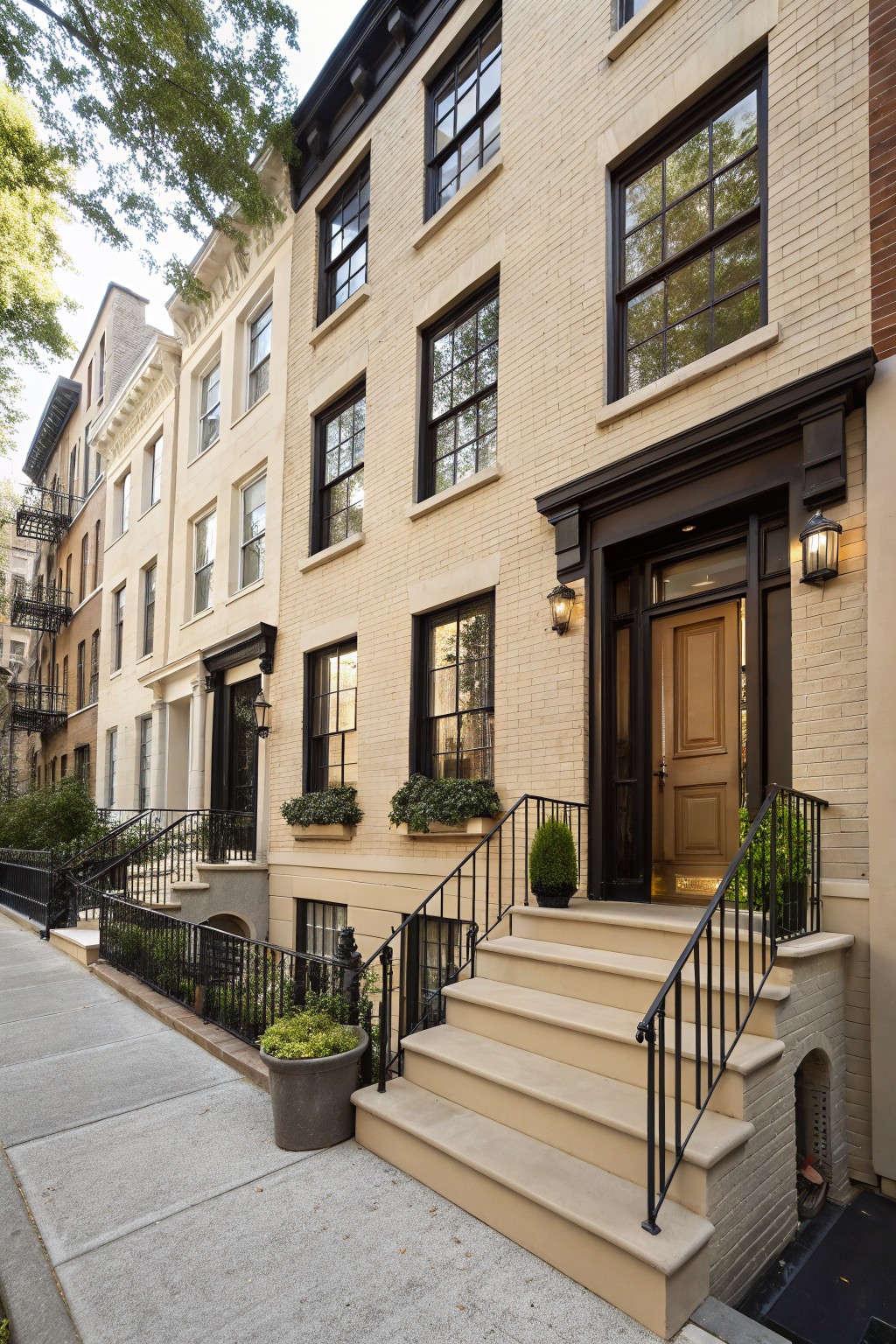 Cream painted brick townhouse exterior with black window frames and trim, wooden front door, beige stone steps with black iron railings, lanterns, and potted plants on a city sidewalk.