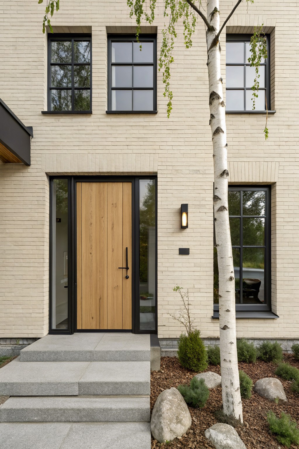 Cream brick house exterior with black-framed windows, a tall wooden front door with sidelight glass panel, concrete steps, wall light, and a birch tree with shrubs and rocks in the front yard.
