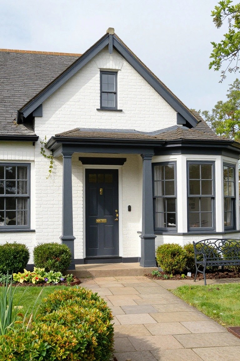 Front exterior of a white brick cottage with dark gray trim on gabled roof edges, porch columns, windows, and bay window, navy front door with brass knocker, slate roof, potted plants, shrubs, bench, and stone path in landscaped yard.