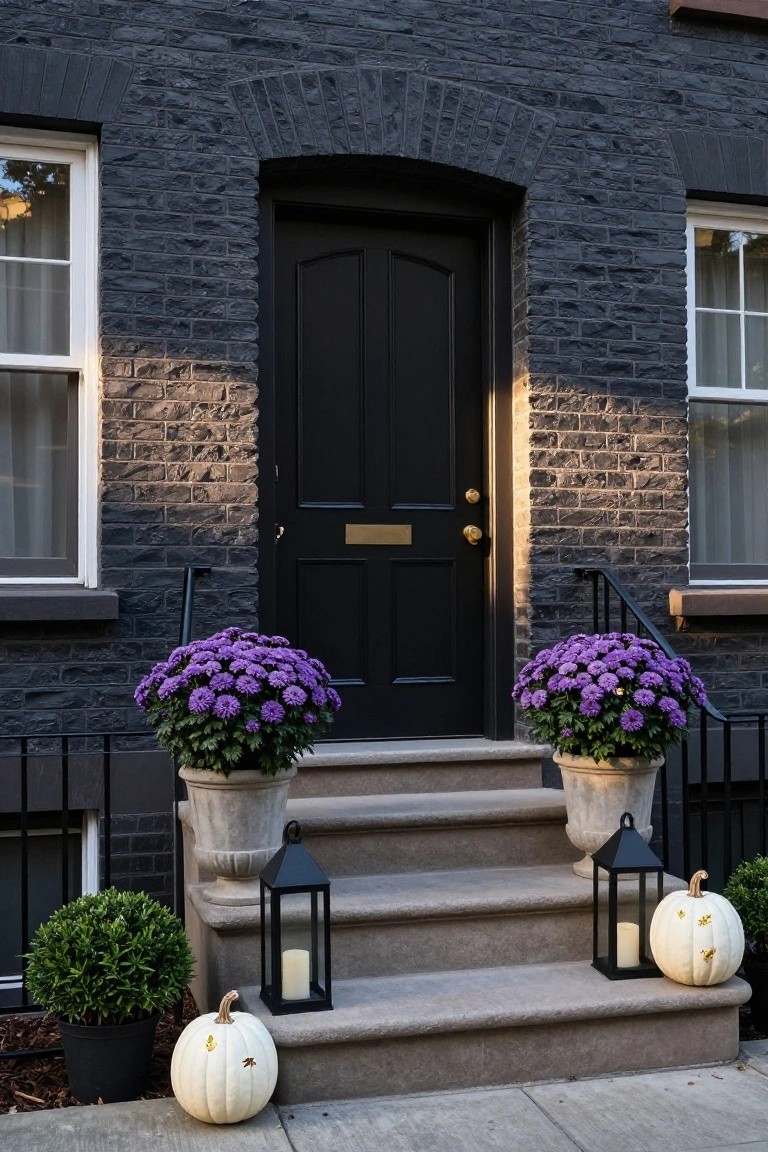 Dark brick townhouse exterior with arched black front door, purple chrysanthemum plants in urns flanking stone steps, white pumpkins with gold designs, black metal lanterns holding candles, and potted boxwood shrubs.