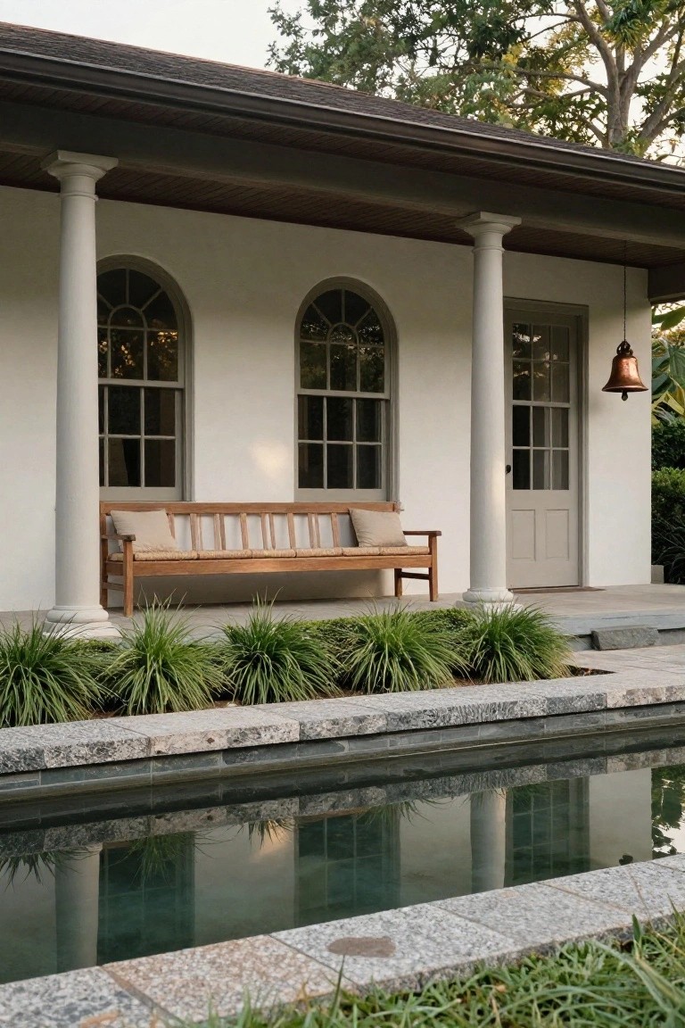 White stucco porch with fluted columns, two arched multipane windows, gray door with brass bell, teak bench with beige cushions and pillows, flanked by grasses and a granite-edged reflecting pool.