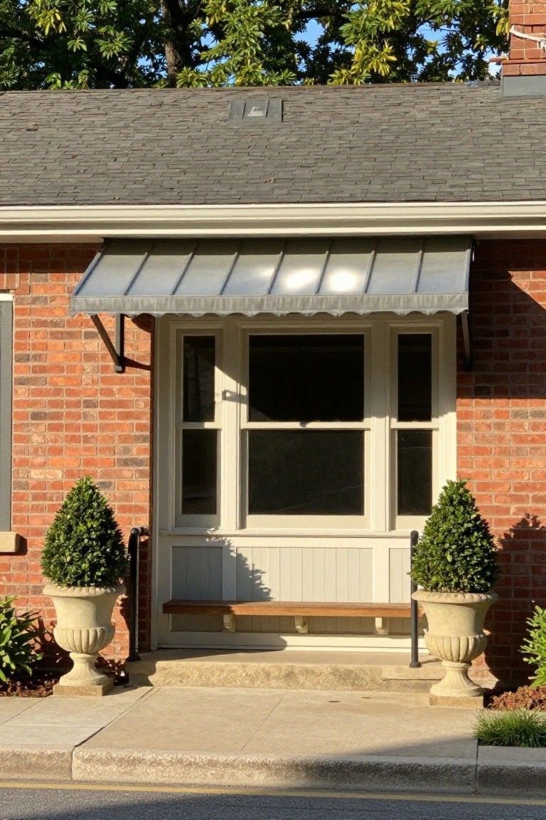 Brick house front entry with gray metal awning over a white door and sidelights, flanked by potted boxwood topiaries in urns, wooden bench below, and stone steps leading to the sidewalk.