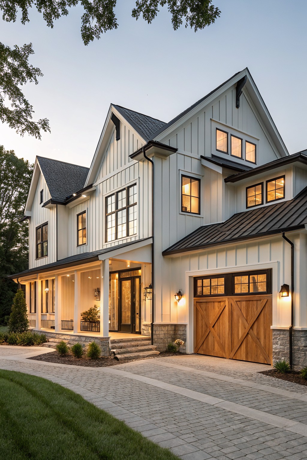 Three-story modern farmhouse exterior with white board-and-batten siding, black metal roofs, black-framed windows, covered front porch, wooden barn-style garage doors, stone accents, pathway, and lawn at dusk.