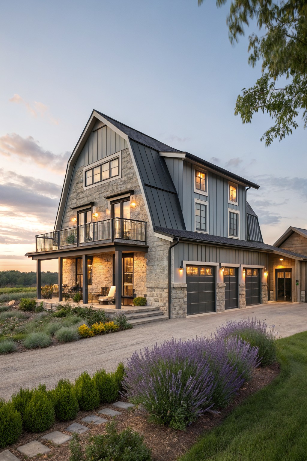 Three-story modern farmhouse with gambrel roof, black metal roofing, gray vertical siding, stone base and accents, wraparound balcony, attached three-car garage, and gravel driveway with lavender plantings at dusk.
