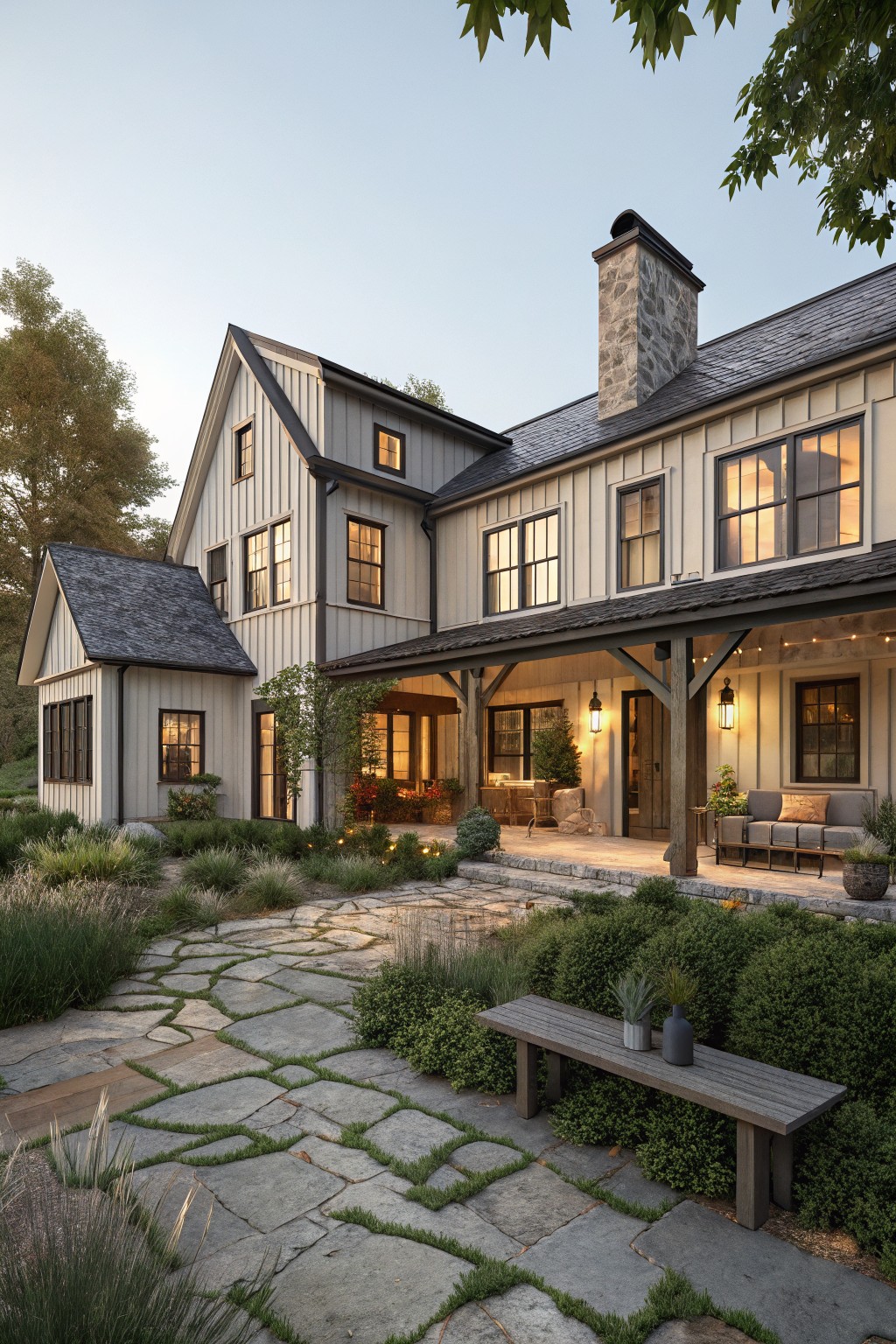 Modern farmhouse house exterior with light board-and-batten siding, dark sloped roofs, stone chimney, deep covered porch with sofa, lanterns, string lights and plants, leading to a stone pathway with bench and ornamental grasses.