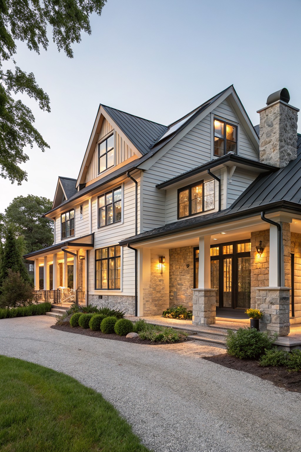 Three-story modern farmhouse house exterior featuring light gray siding, black metal roof with gables, tall stone chimney, wraparound porch with stone columns, black-framed windows, gravel driveway, shrubs, and trees at dusk.
