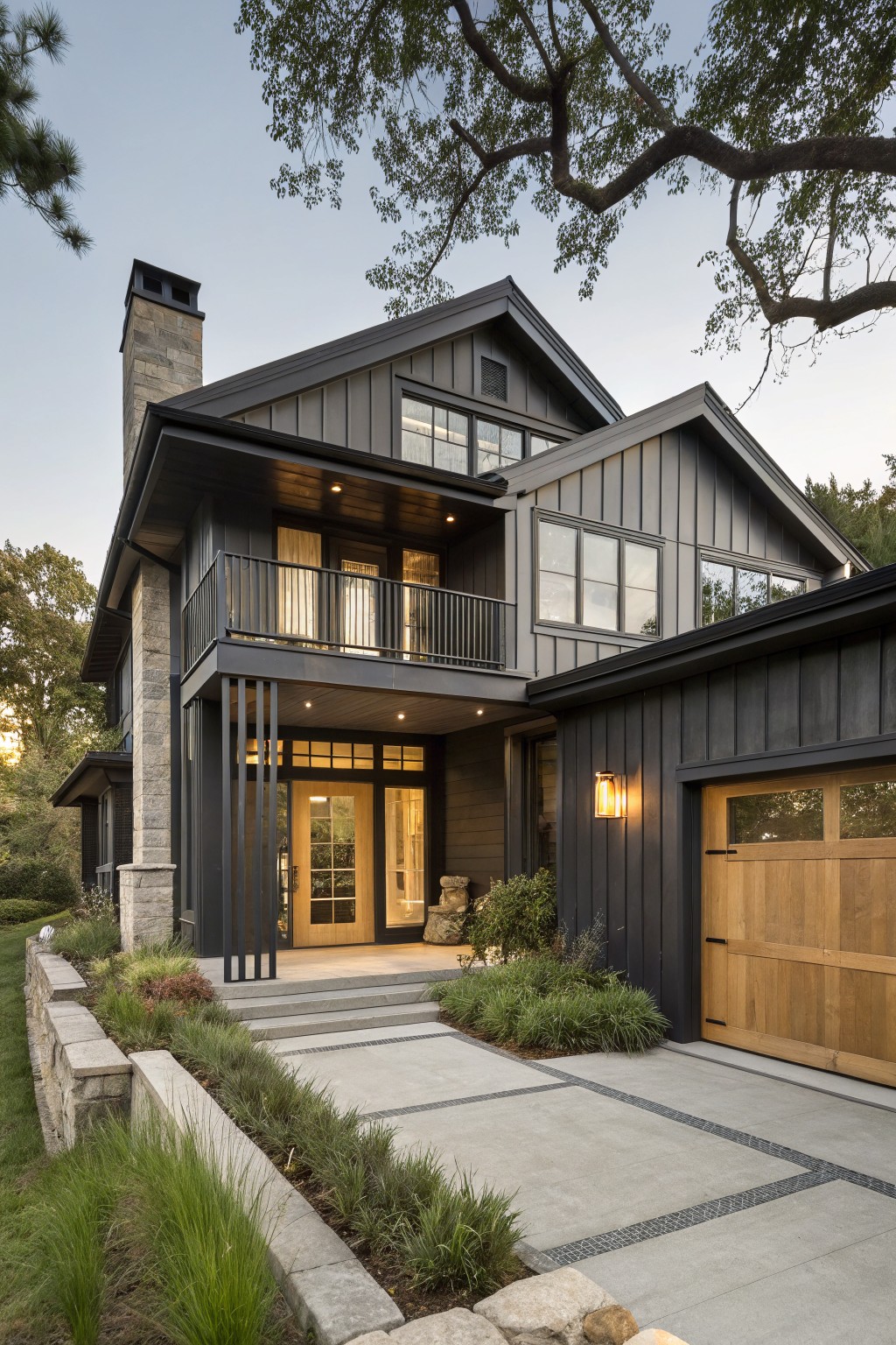 Two-story modern farmhouse exterior with black board and batten siding, stone chimney, metal balcony, covered entry with wood doors, wood garage door, concrete pathway, stone retaining wall, and surrounding plants and trees at dusk.
