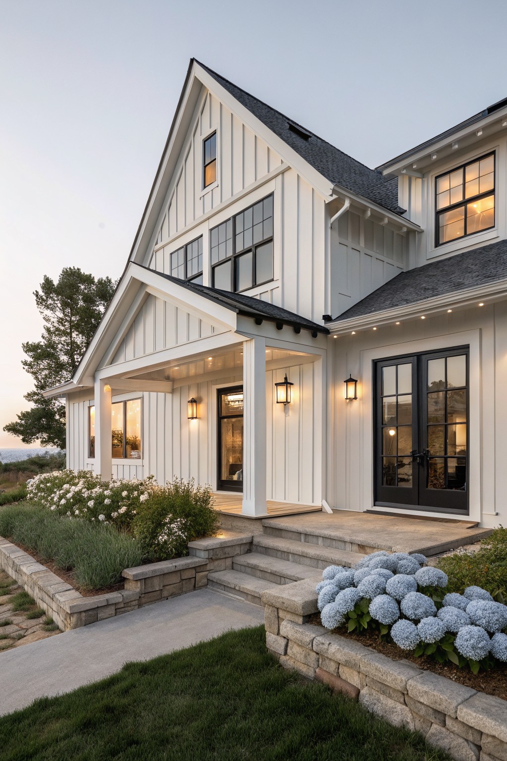 White board and batten sided modern farmhouse exterior with gabled roof, covered front porch, black framed windows and double doors, stone steps and retaining wall with hydrangeas and shrubs, at dusk.