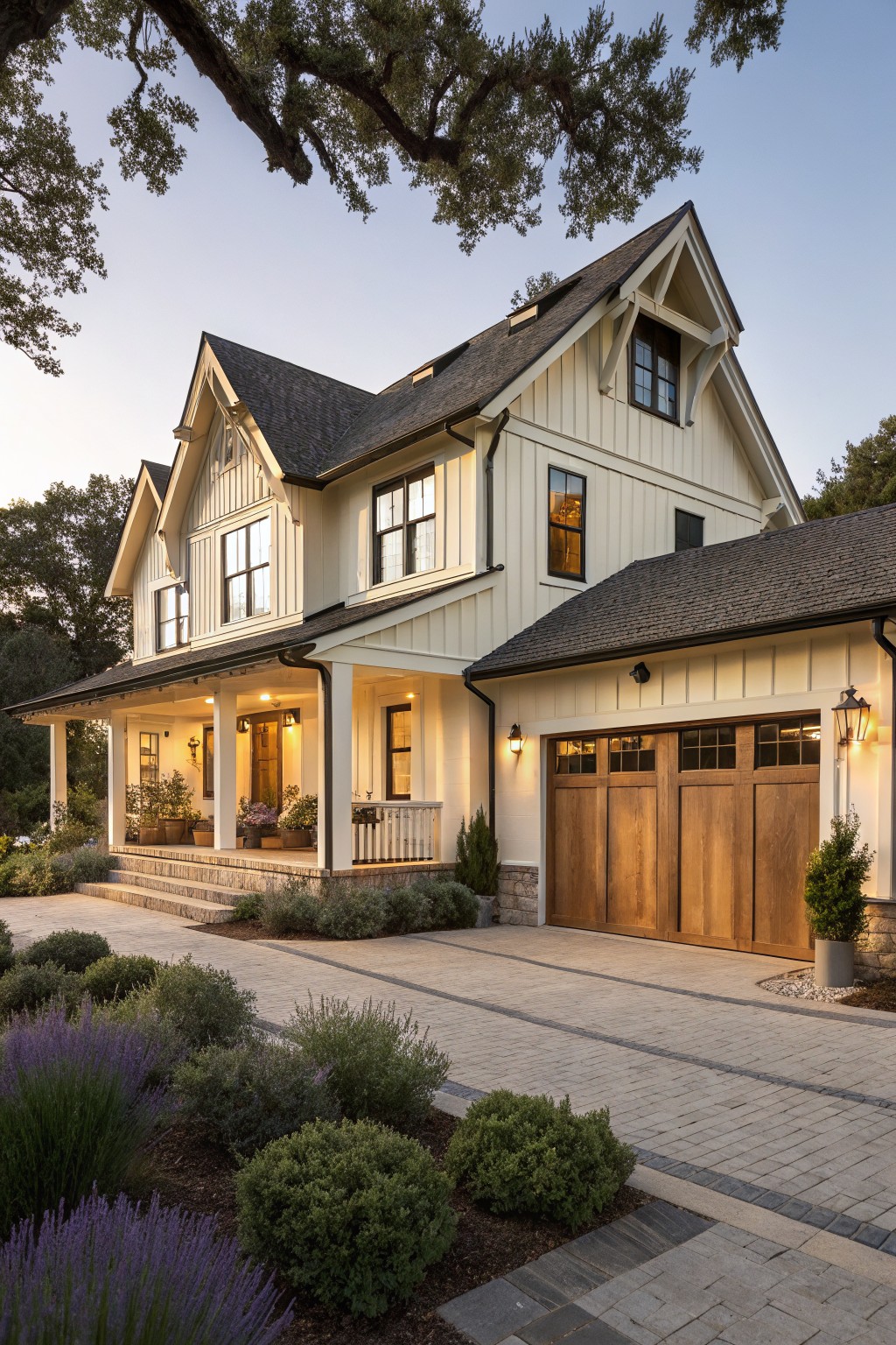 Two-story cream board and batten farmhouse exterior at dusk with gabled roofs, covered front porch, attached garage with wood doors, and paved walkway amid shrubs and lavender plants.
