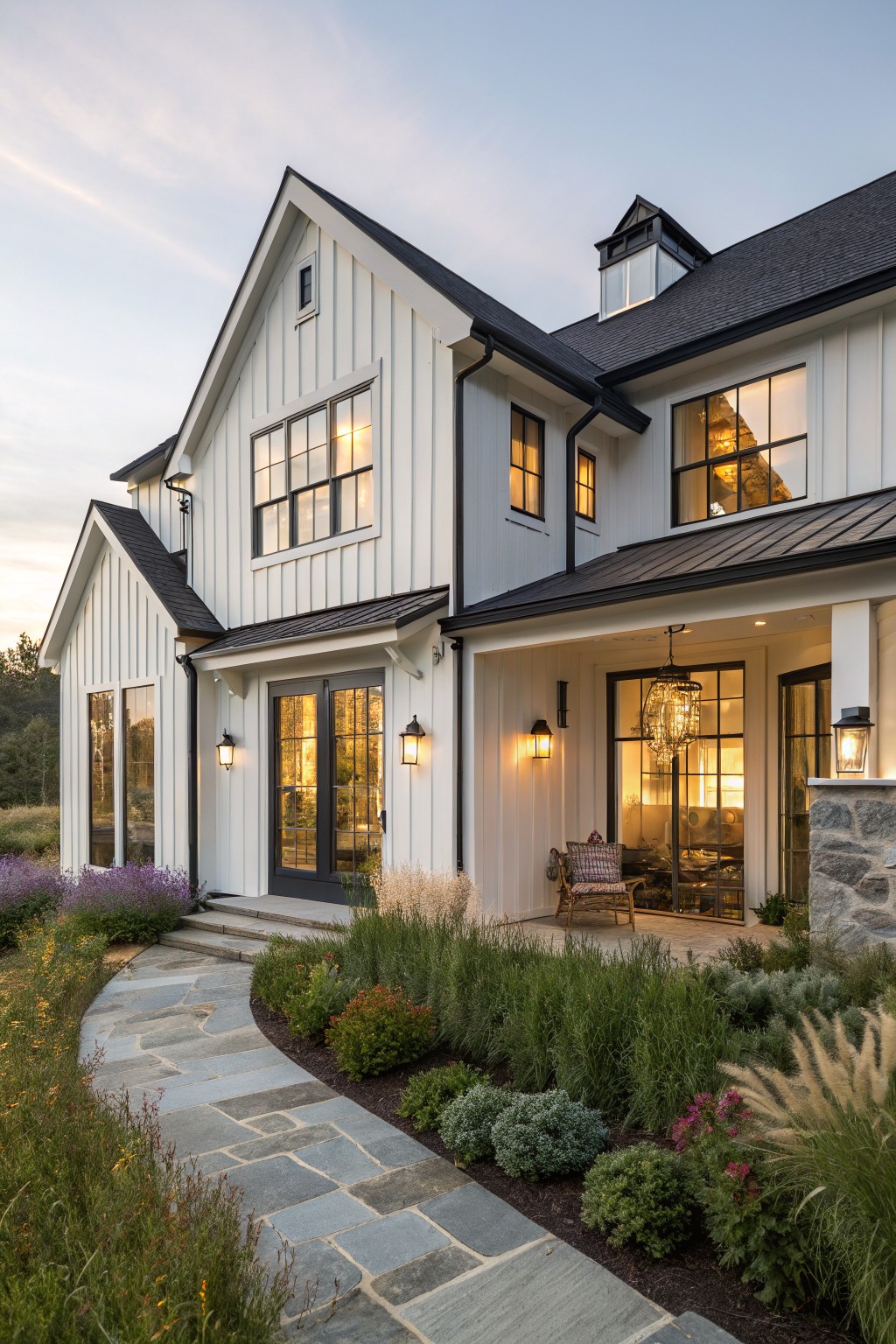 White board and batten house exterior with black metal roof, gabled rooflines, large black-framed windows and doors, covered front porch, stone steps, and surrounding landscaping with grasses and perennials at dusk.