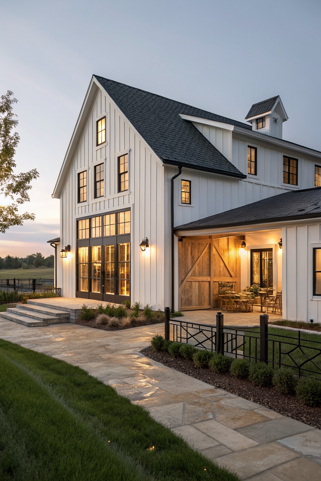 White board and batten sided two-story modern farmhouse exterior at dusk with steeply pitched dark roof, large black-framed windows, sliding wooden barn door under covered porch, stone steps and pathway, and surrounding landscaping.