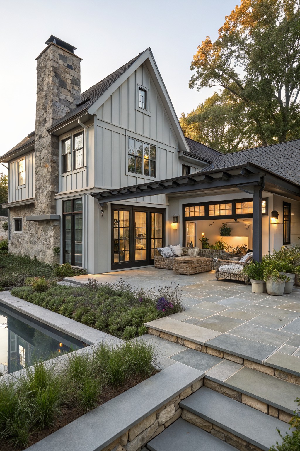 Rear and side exterior view of a two-story modern farmhouse with light gray board and batten siding, tall stacked stone chimney, black metal pergola covering a patio with wicker furniture, stone steps leading to an in-ground pool, and low plantings along the edges.