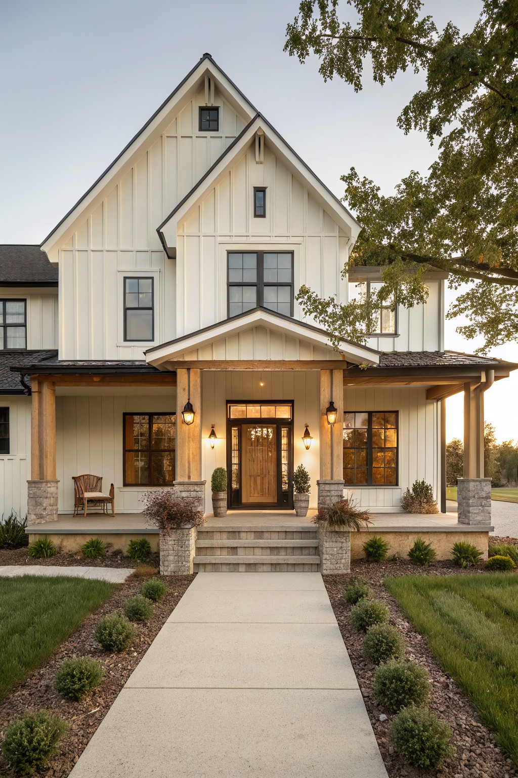 Two-story house exterior with white board and batten siding, black-framed windows, covered front porch supported by wooden posts on stone bases, lanterns, potted plants, steps, and landscaped yard with shrubs and grass pathway at sunset.