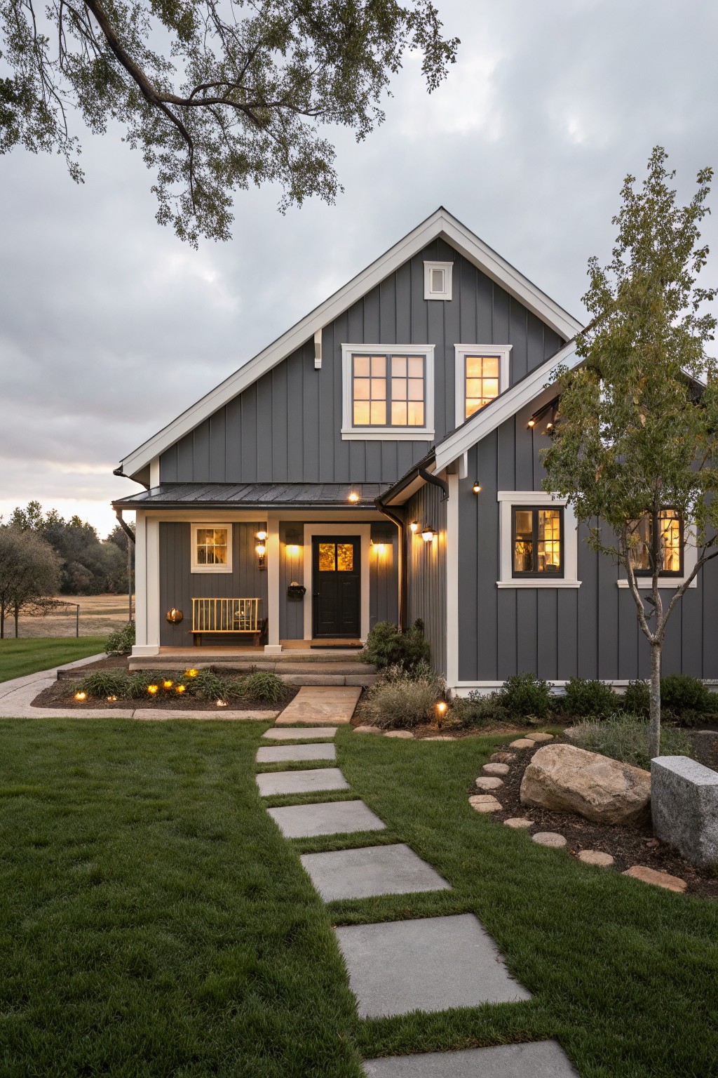 Front exterior view of a modern farmhouse house with dark gray board and batten siding, white trim accents, gabled roofline, covered porch with bench and lights, stone pathway, landscaped yard, and trees at dusk.