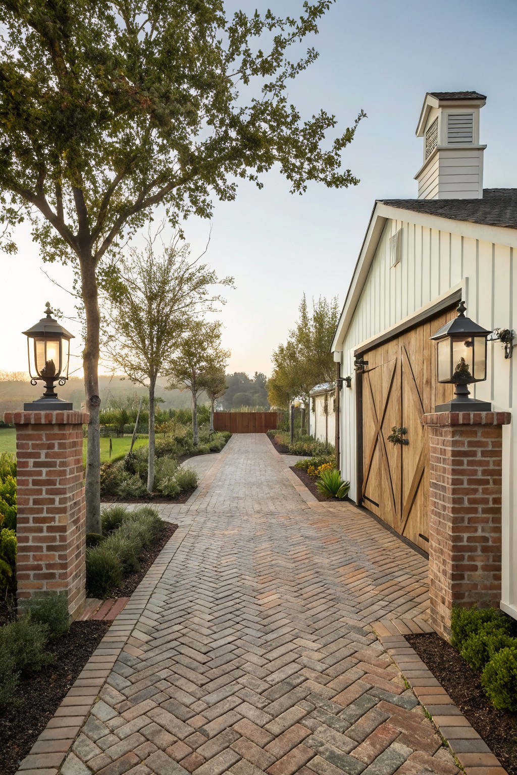 Herringbone-patterned brick paver pathway flanked by brick pillars topped with lanterns, leading to large wooden barn doors on a white shiplap building, with landscaped beds, trees, and grass on either side.