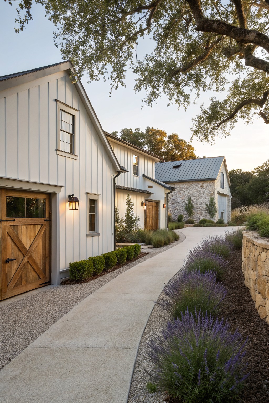 White board-and-batten garage with wooden X-braced double doors, curved concrete driveway edged by lavender plants, gravel margins, boxwood shrubs, and stone retaining wall, with a second stone barn structure and oak trees in the background at dusk.
