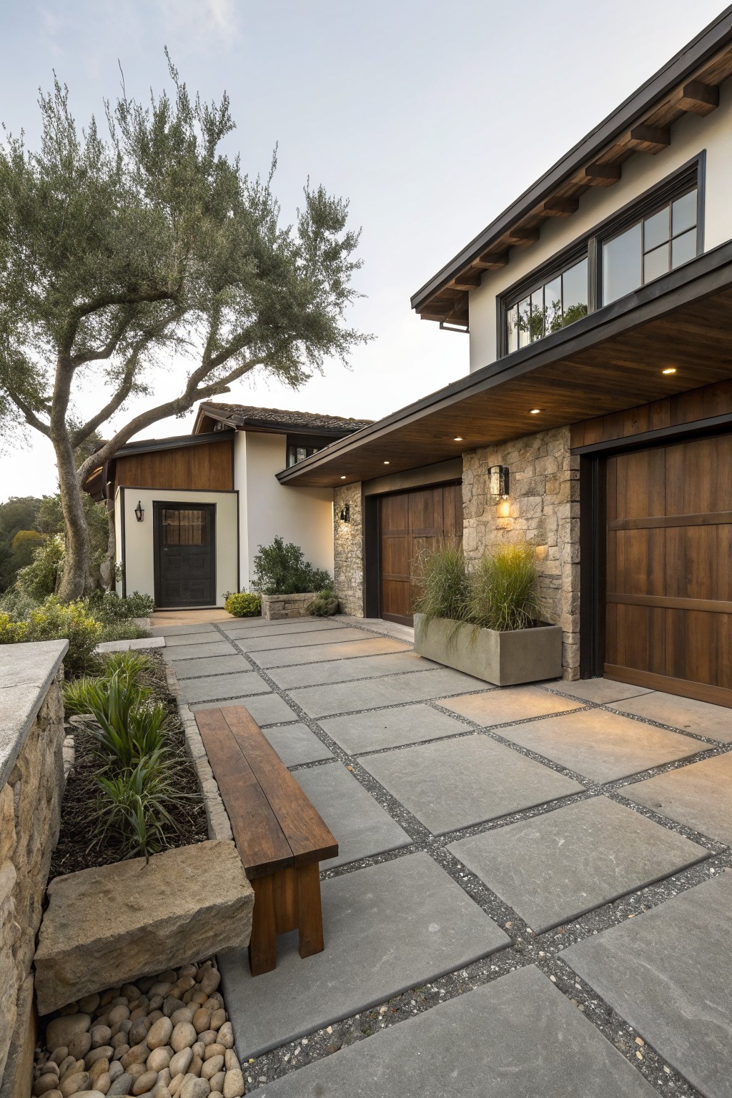 Side view of a modern house exterior showing a driveway paved with large gray rectangular pavers edged with pebbles and plants, a wooden bench, stone retaining wall, wooden garage doors, and an olive tree.