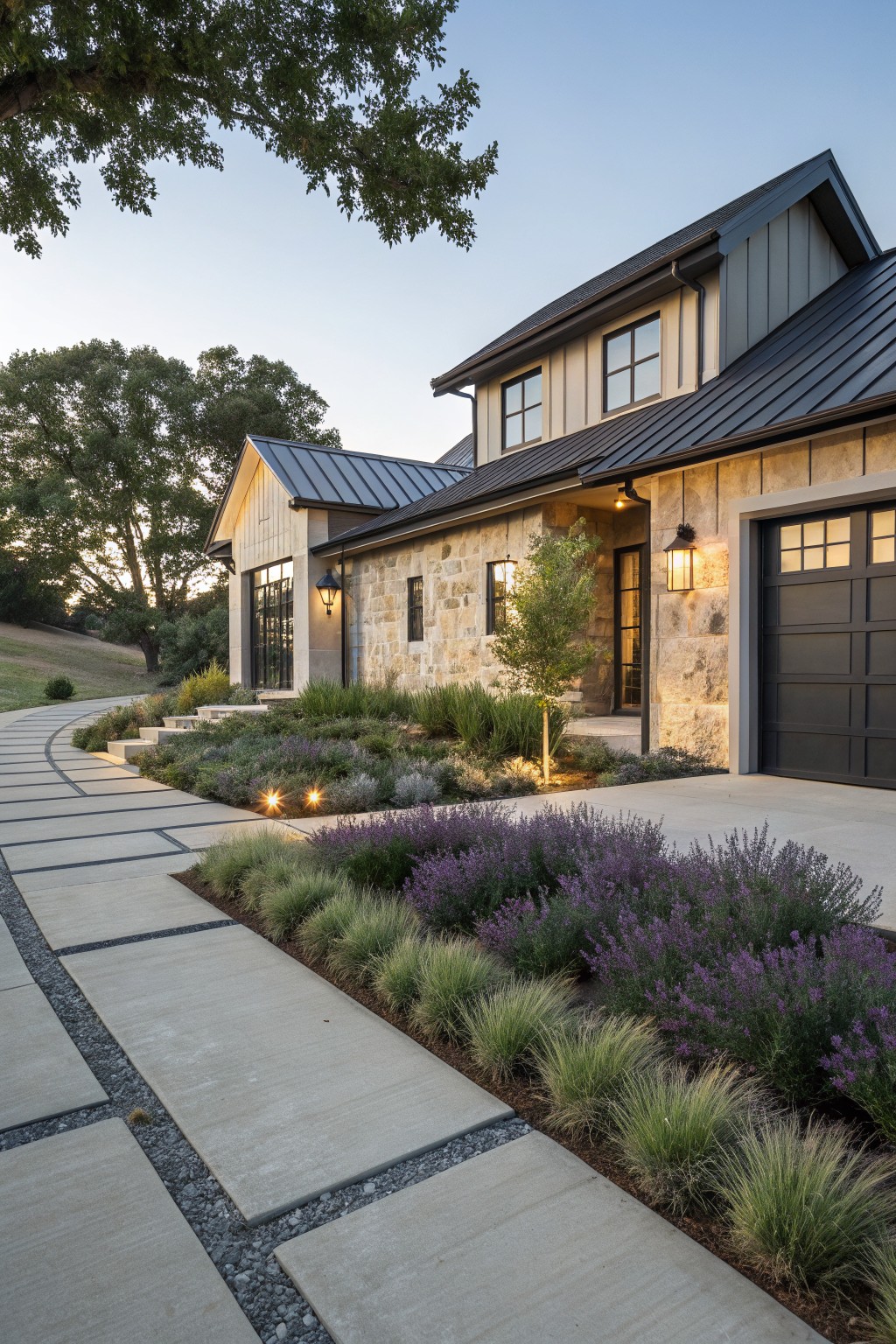 Modern farmhouse garage exterior with curved concrete slab pathway edged by lavender shrubs and ornamental grasses, stone walls, black metal roof, and pathway lights.