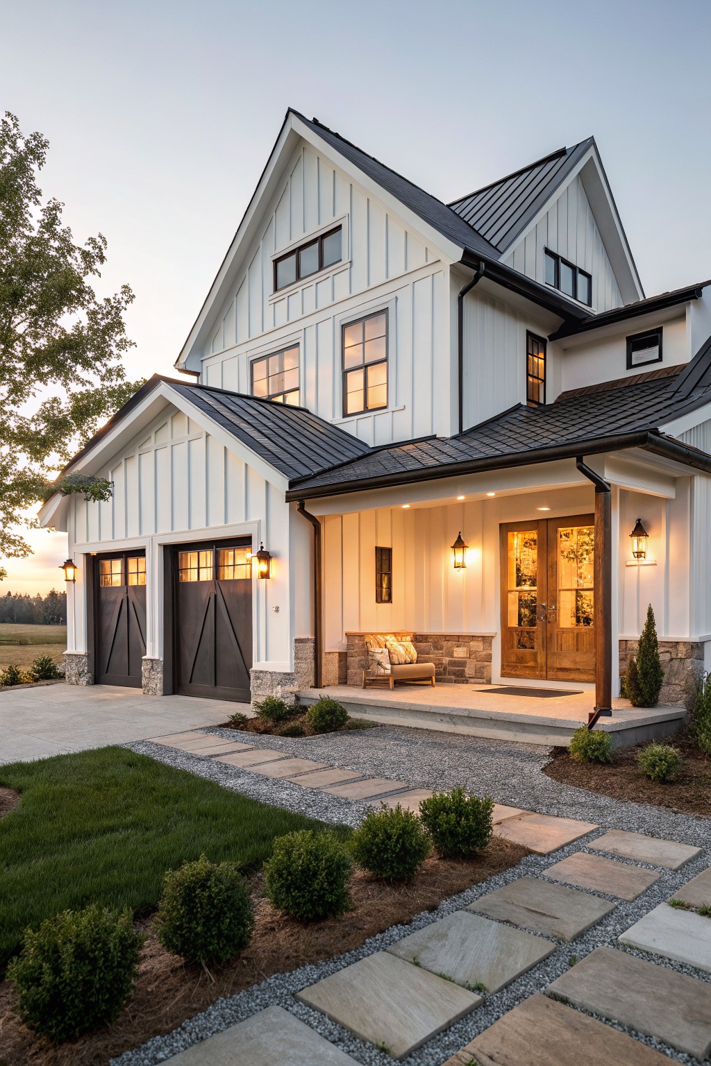 White board-and-batten farmhouse with black metal roof, dark garage doors, covered front porch with lanterns, stone base, and gravel path with boxwoods in front yard at sunset.