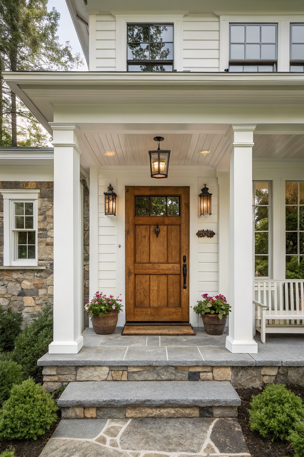 White clapboard farmhouse exterior with covered porch, wooden paneled front door, black lanterns, stone steps, potted flowers, and white bench.