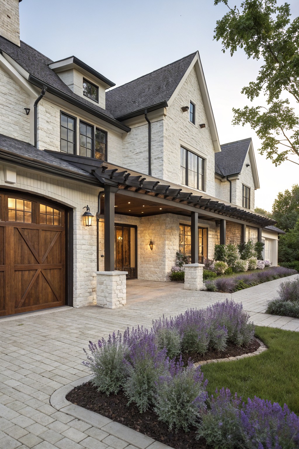 White brick modern farmhouse house exterior with black pergola-covered porch spanning over wooden garage door and stone entry, lavender shrubs along paver driveway and lawn edge.