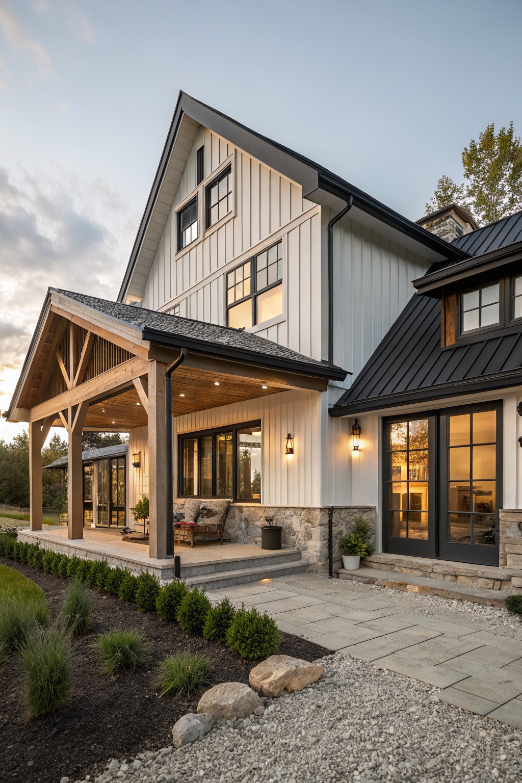 Side view of a modern farmhouse house featuring white vertical siding, black metal roof sections, a covered gabled porch with exposed wooden beams and posts, stone foundation details, a stone pathway, low shrubs, and gravel mulch.