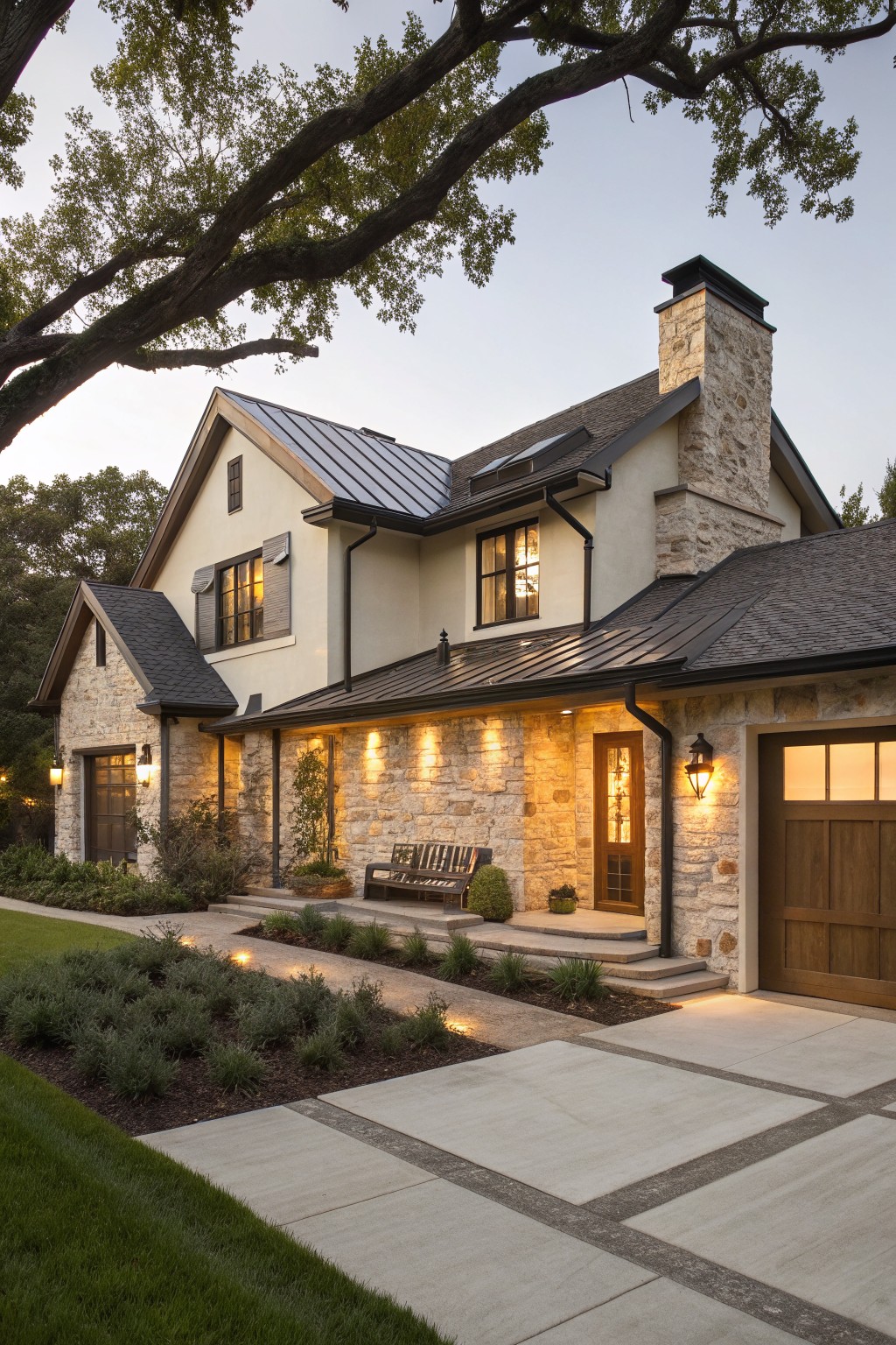 A modern farmhouse exterior with beige stucco walls, limestone block accents on chimney and garage, dark metal roof, wood garage door, lit entry steps, and low plantings along a concrete path under large trees at dusk.
