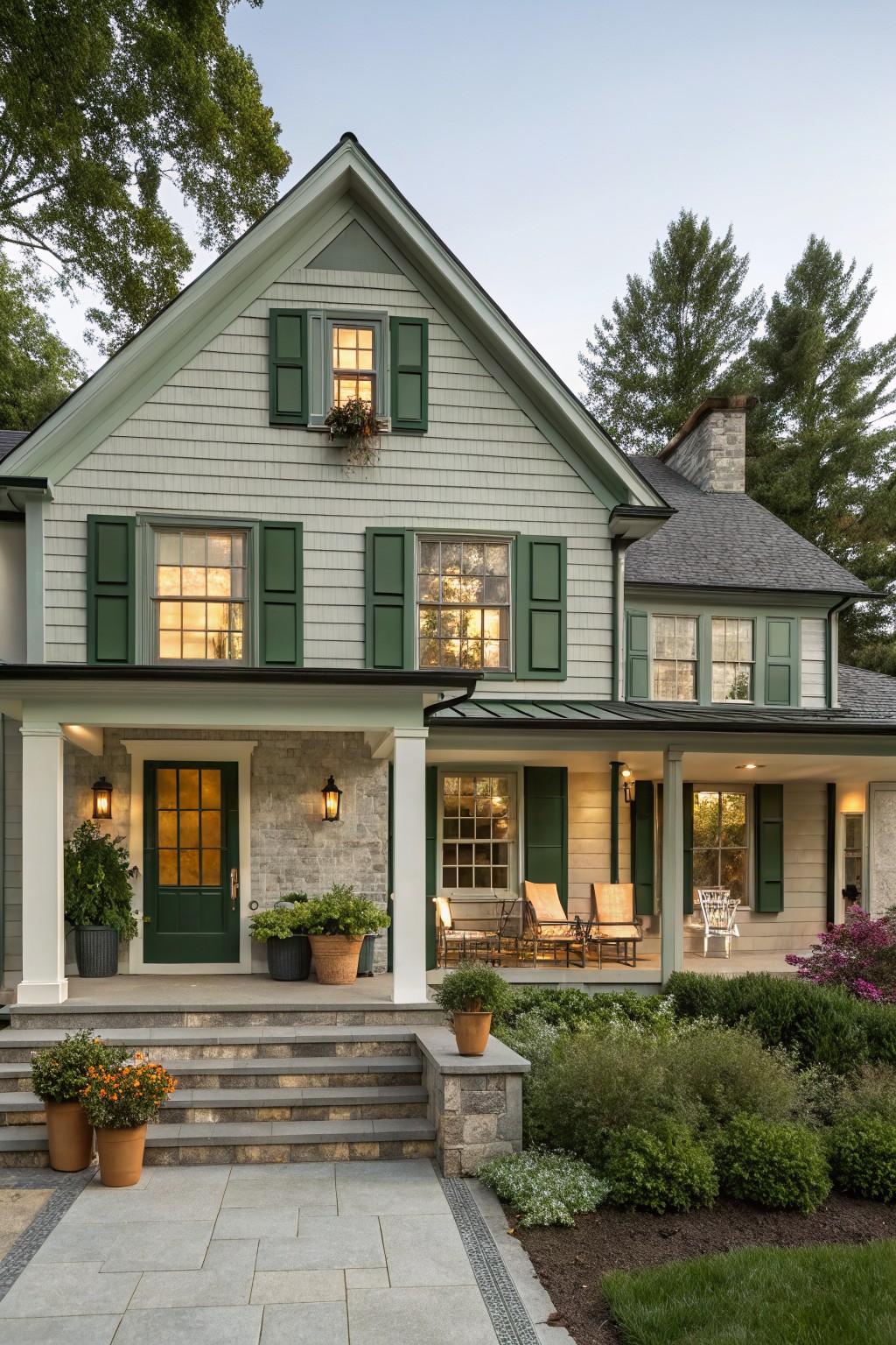 Front view of a two-story house with pale green shingle siding, green shutters on multipane windows, covered porch with green door and lanterns, stone steps, potted plants, and landscaped shrubs.