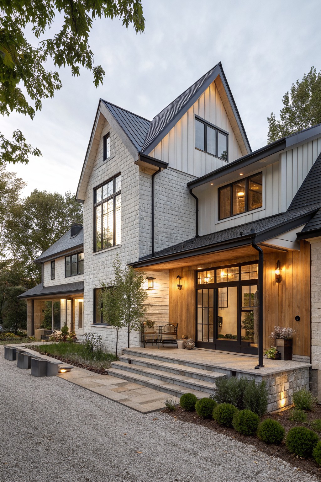 Two-story modern farmhouse house exterior featuring light gray stone lower walls, white board-and-batten siding, black metal gabled roof, large black-framed windows, covered wooden entry porch with stone steps, gravel driveway, and surrounding trees and shrubs.